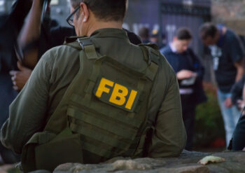 Caucasian male FBI agent wearing glasses in a green bulletproof vest with FBI logo seen from behind on the investigation spot.